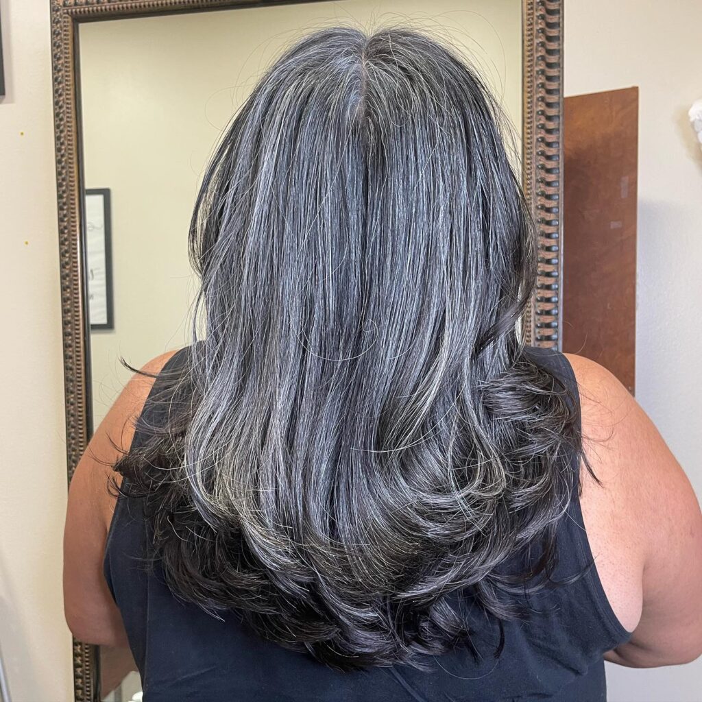 A woman with thick salt and pepper hair styled in long layers with the ends flipped outward.