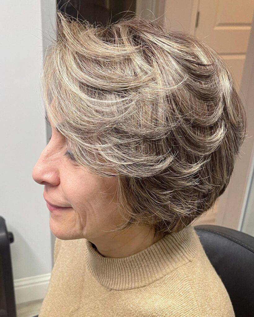 A woman with thick ash blonde highlighted hair styled in a voluminous feathered bob with swept back layers.