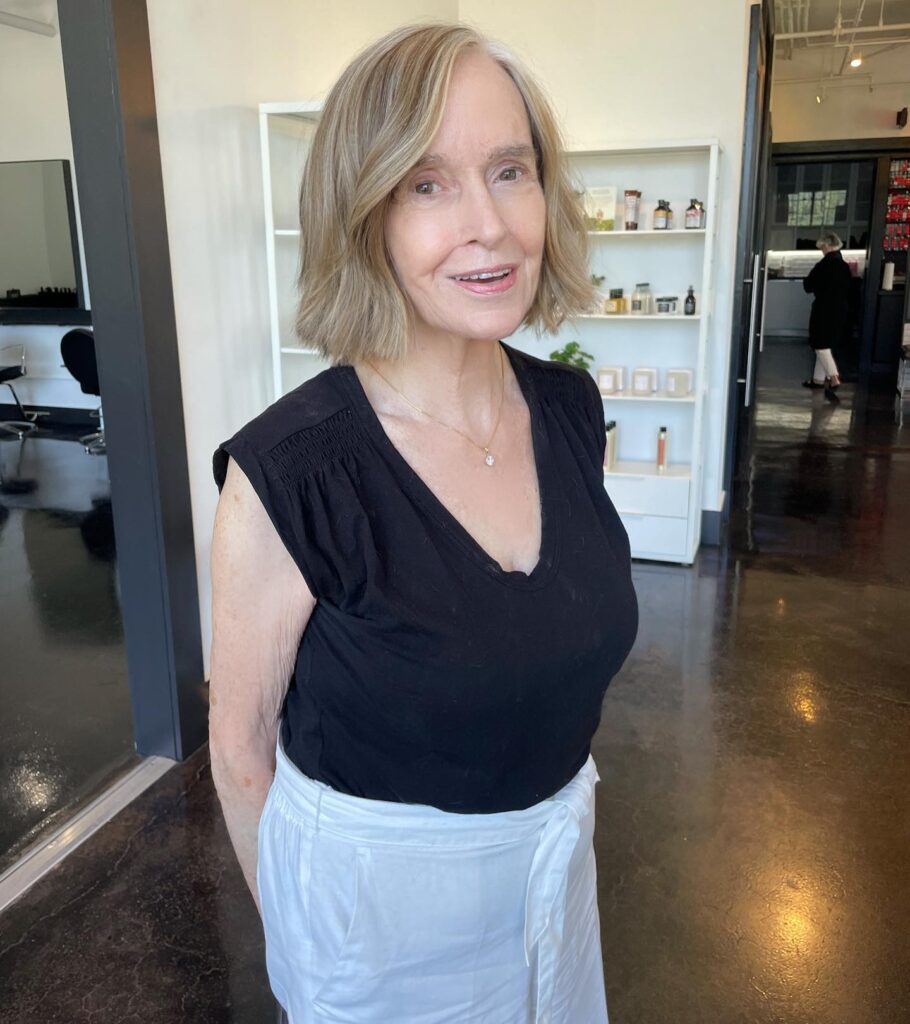 A smiling woman over 70 wearing a black top and white pants with a honey blonde chin length bob and soft waves.