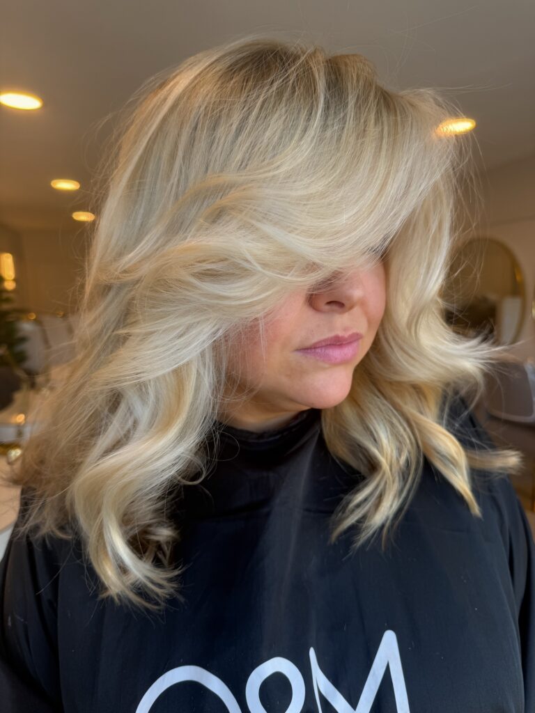 A woman with thick shoulder length creamy blonde hair styled in voluminous glamorous waves with a heavy side sweep.