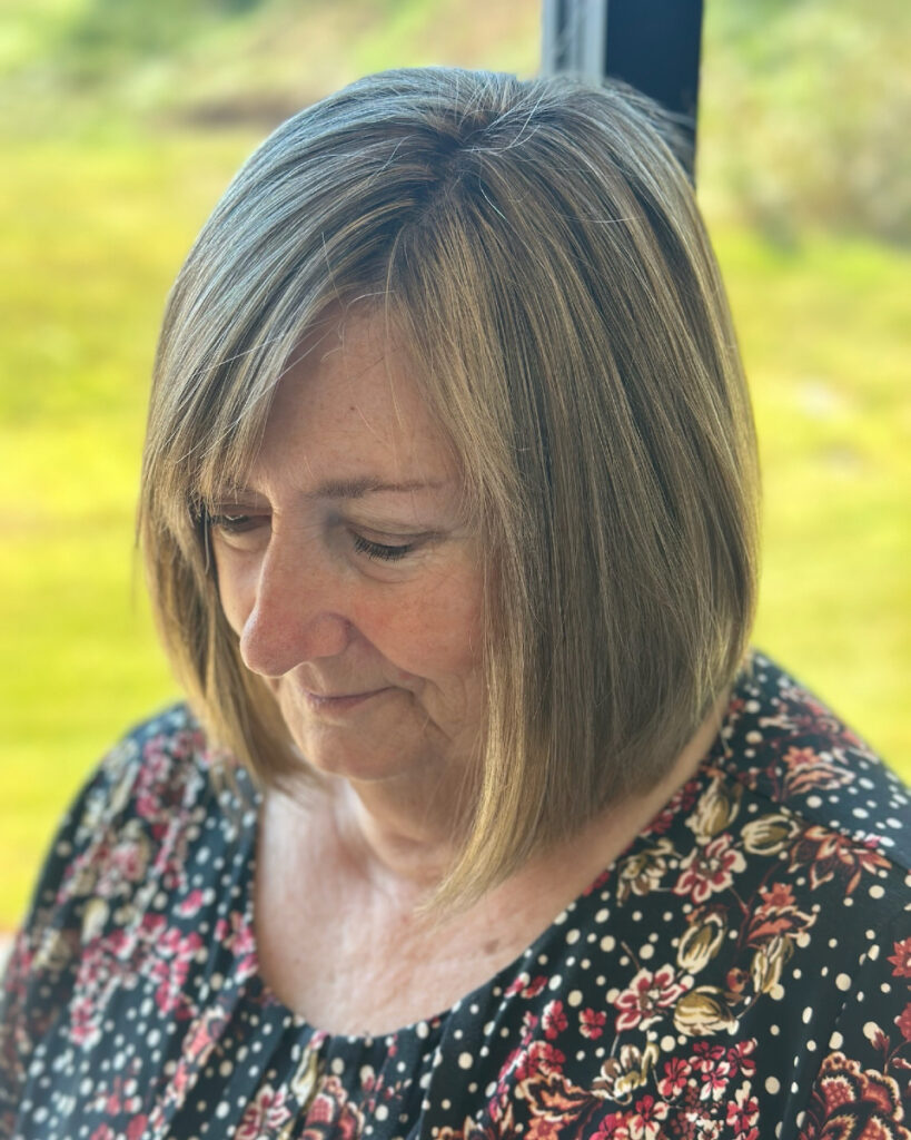 A woman with a chin length angled ash blonde bob looking downward in a garden setting.