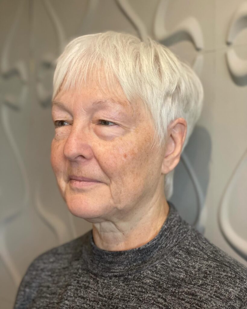 A woman with a short, smooth, icy white pixie cut and soft bangs is looking toward the side.