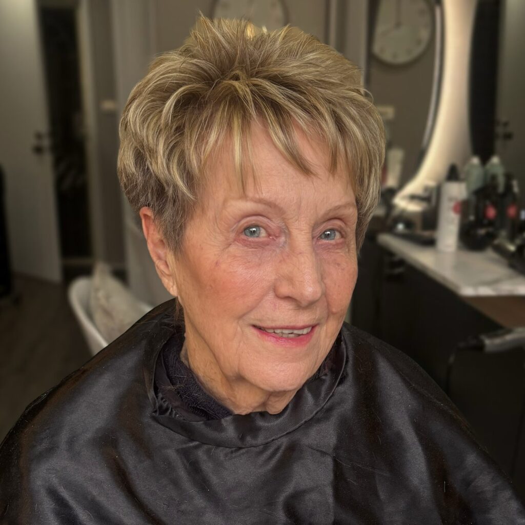 A woman over 70 wearing a textured honey blonde pixie haircut with voluminous layers on top and a soft fringe.