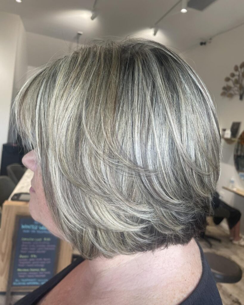 A side profile photograph of a woman with a layered, neck length bob featuring prominent silver ash highlights over a natural grey or light brown base.