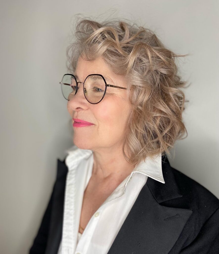 A woman with a voluminous chin length sandy blonde curly bob and black glasses looking to the side.