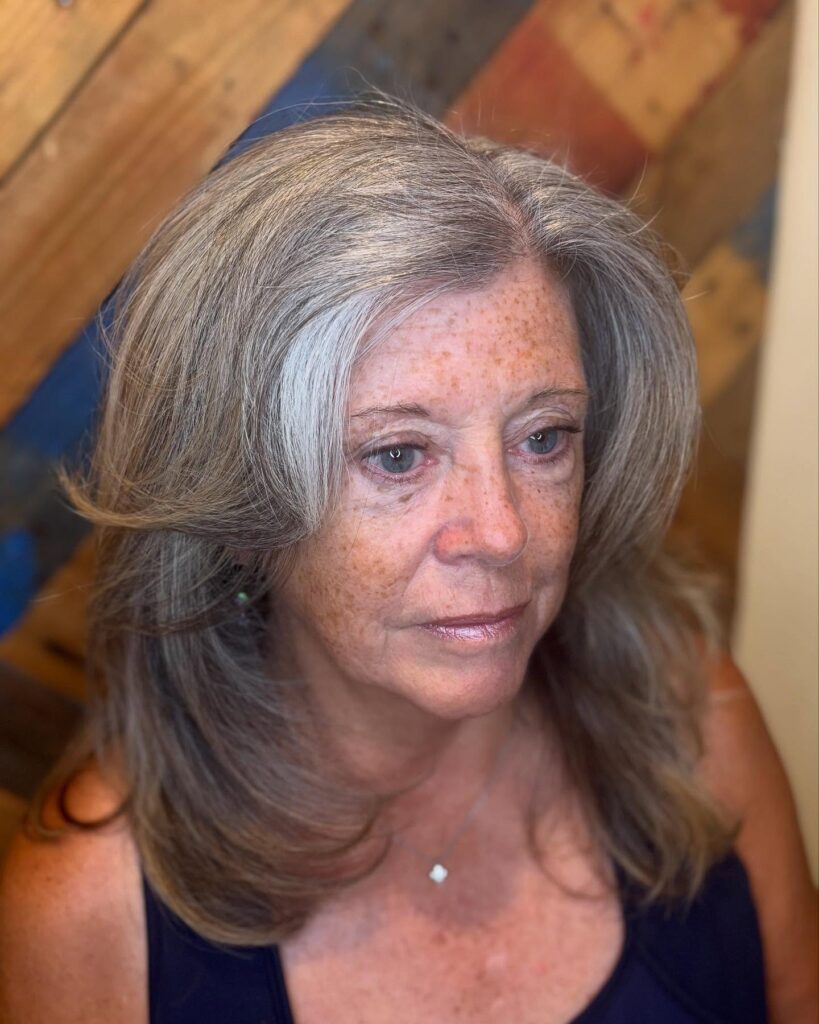 A woman with mid length salt and pepper hair styled with outward sweeping layers and a side part.