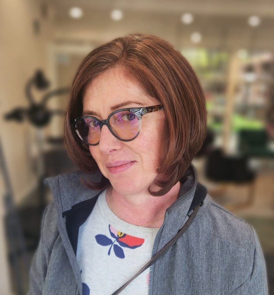 A woman with a sleek chin length mahogany brown bob that curves inward at the ends and black glasses.