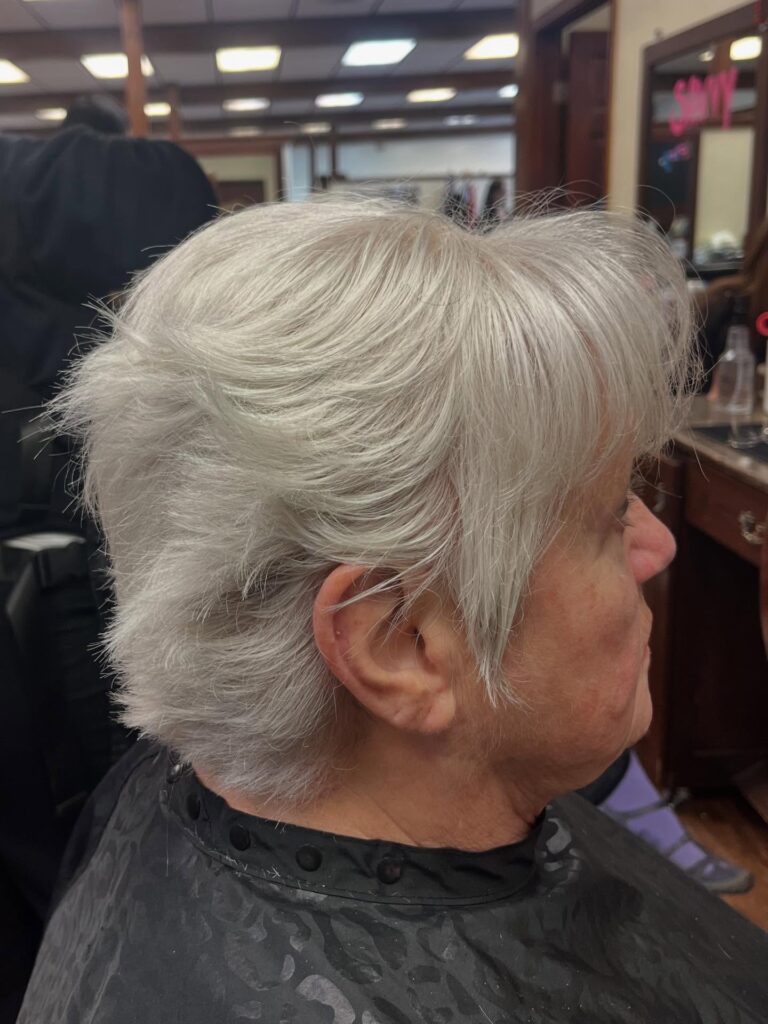 A woman with thick silver hair wears a short feathered pixie cut with soft side swept bangs and textured layers.