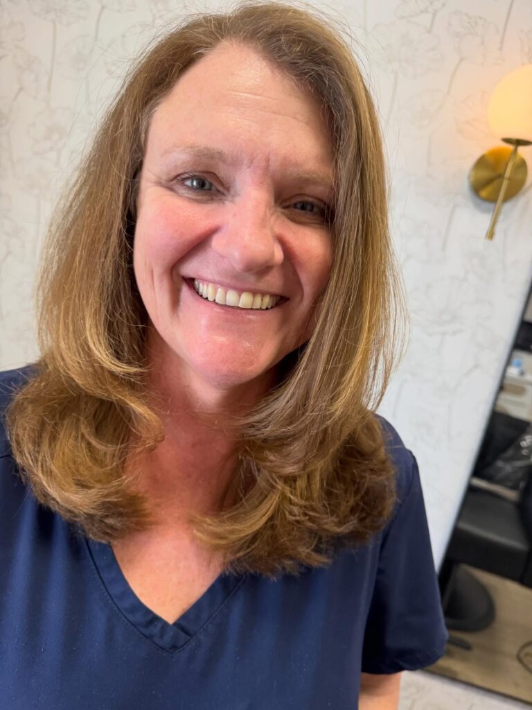 A smiling woman with thick golden brown hair wears a mid length layered cut with ends that curl softly toward her face.