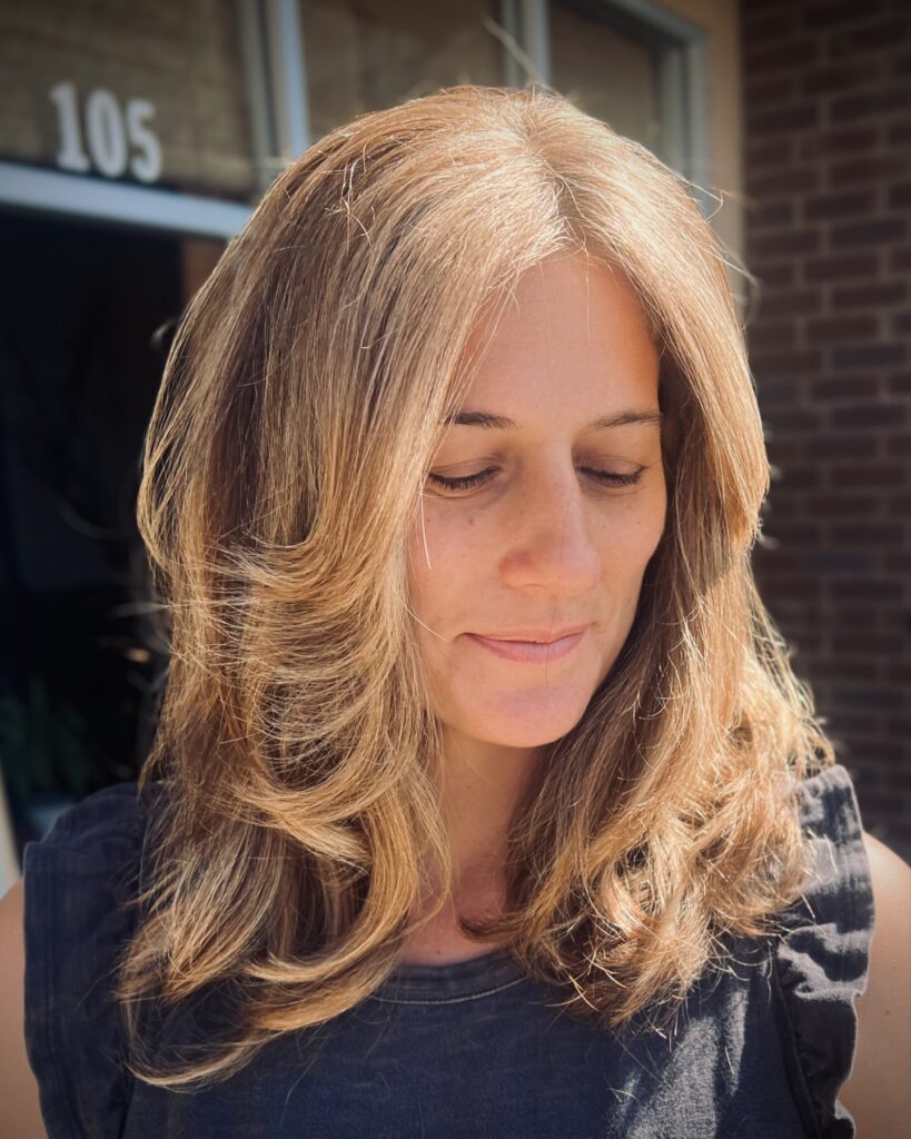 A woman with thick shoulder length honey blonde hair featuring dramatic face framing layers and soft waves.
