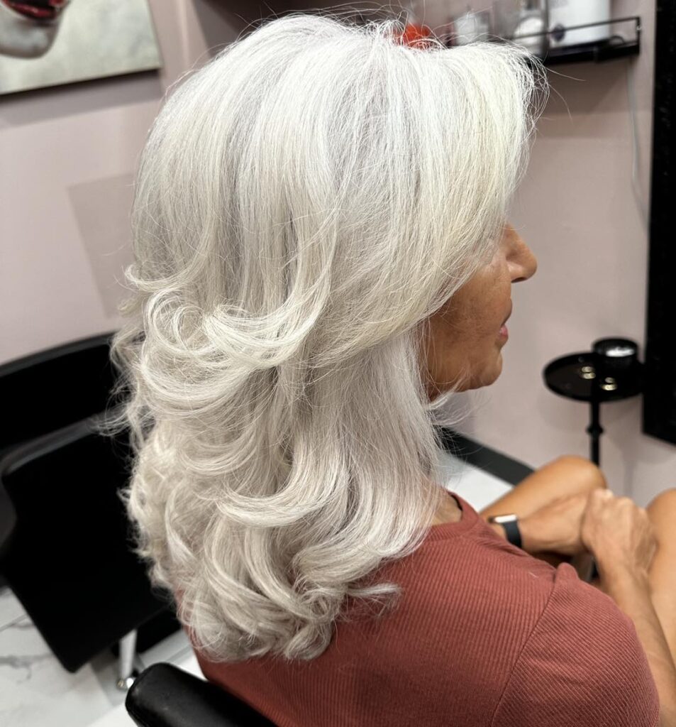 A woman with long thick silver hair styled in voluminous cascading curls and soft layers.