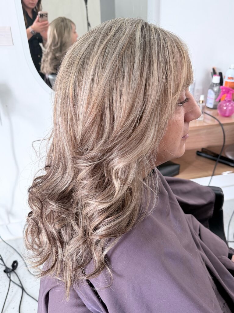 A woman with long thick wavy hair in a honey blonde color featuring soft bangs and layered curls at the ends.