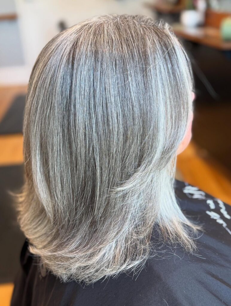 A woman with thick straight silver hair features a shoulder length cut with soft layers and ends that flick outward.