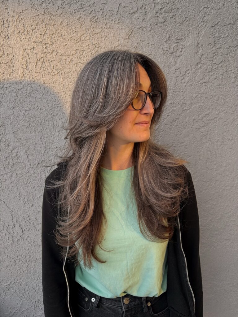 A woman with long thick salt and pepper hair wears a shaggy layered cut with dramatic face framing pieces and wavy ends.