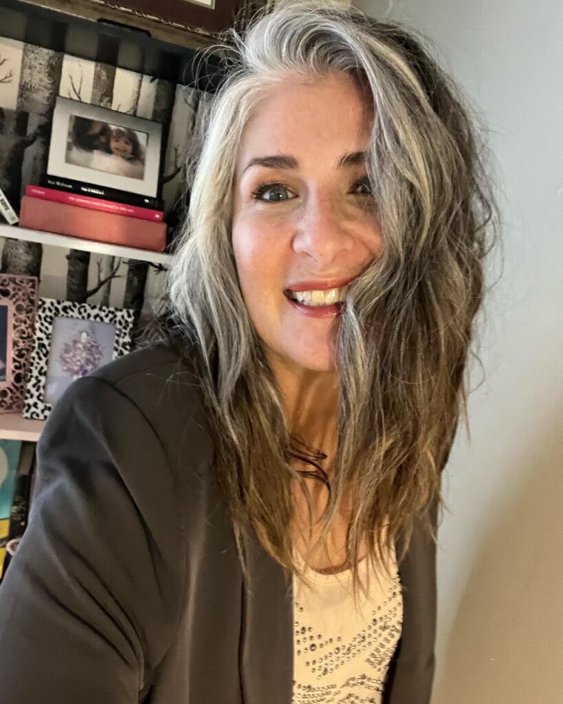 A smiling woman with shoulder length wavy ash brown hair and natural silver highlights.
