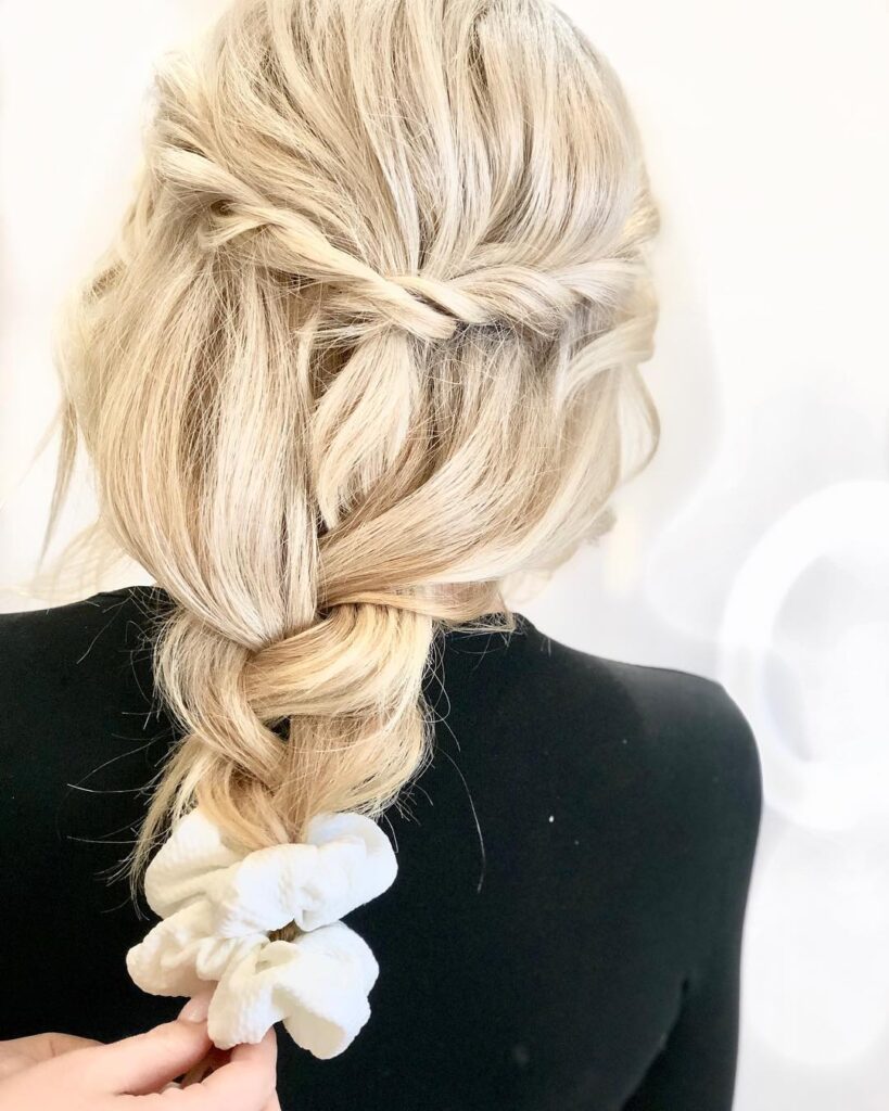 Blonde hair styled in a loose braid that flows into a low ponytail with a scrunchie.