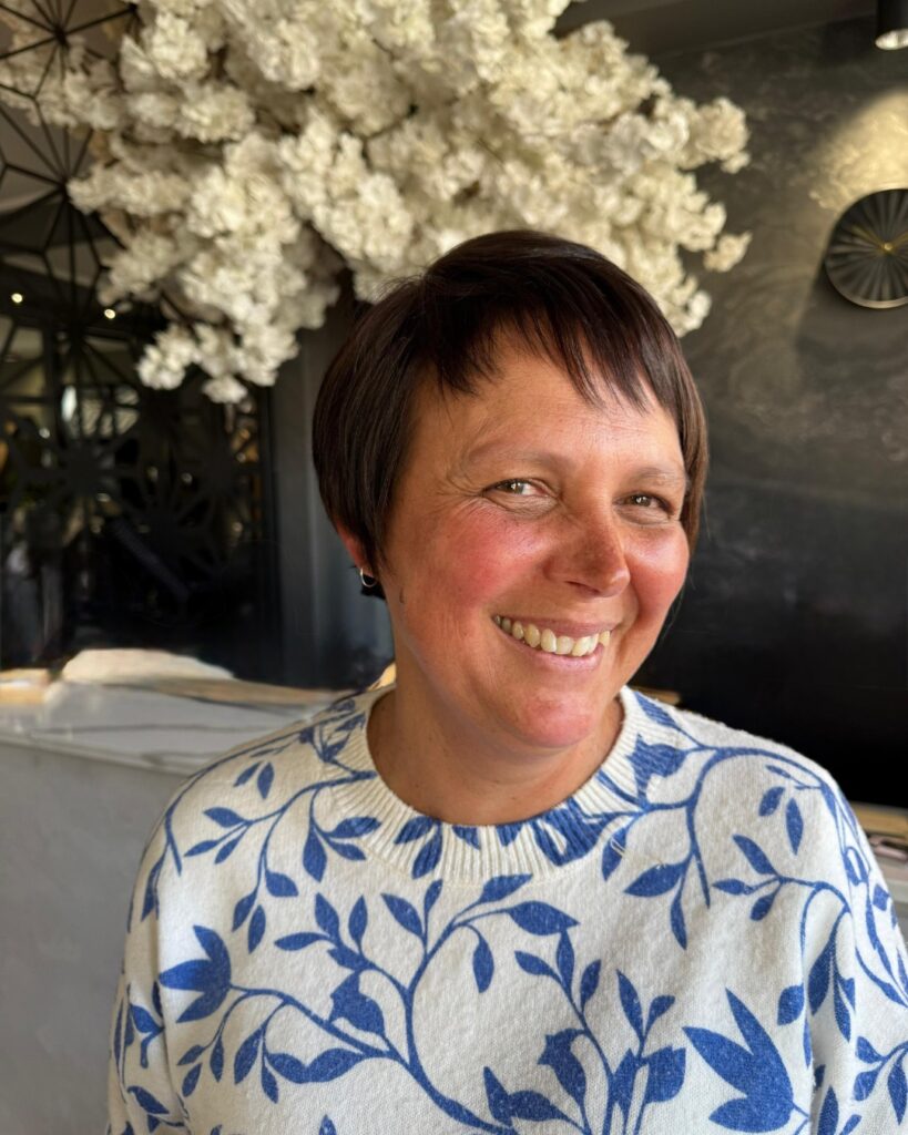 A woman smiling with a short chocolate brown pixie haircut featuring textured layers and a wispy micro fringe.