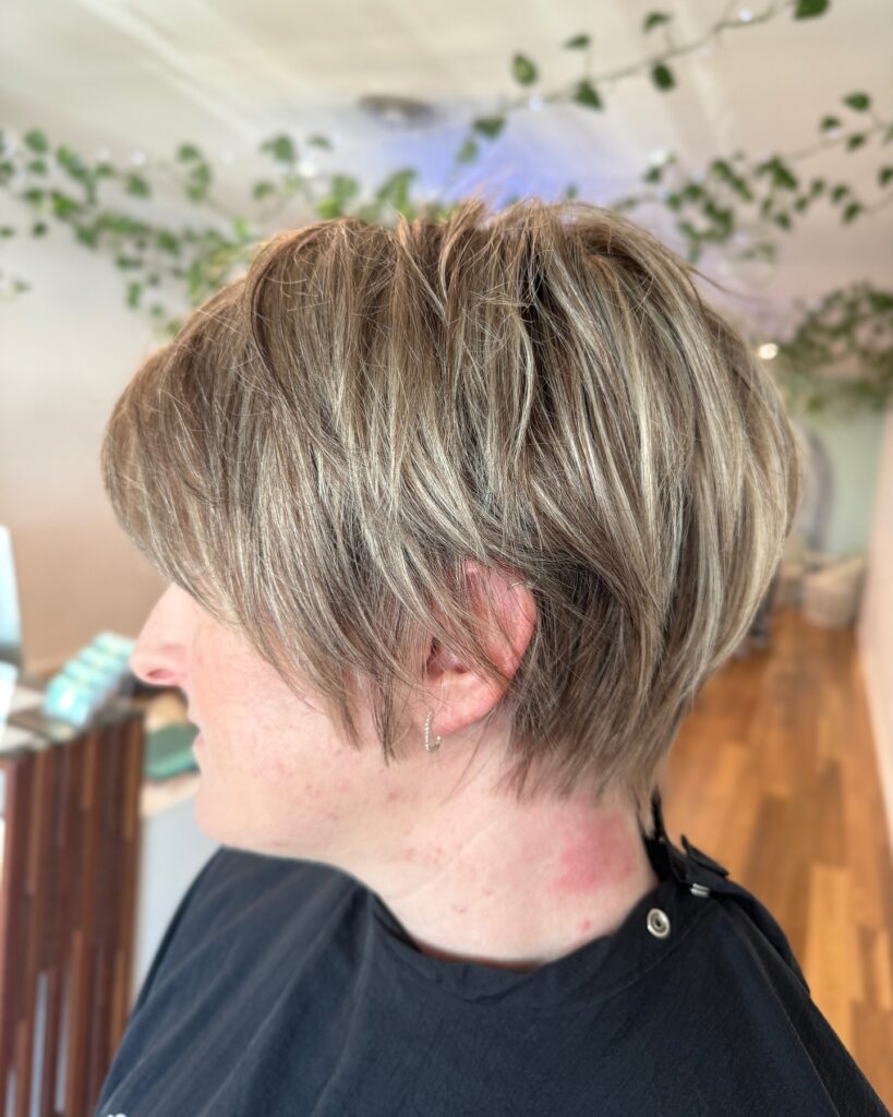 A side profile of a woman showing a textured ash blonde bixie haircut with wispy layers and dark lowlights.