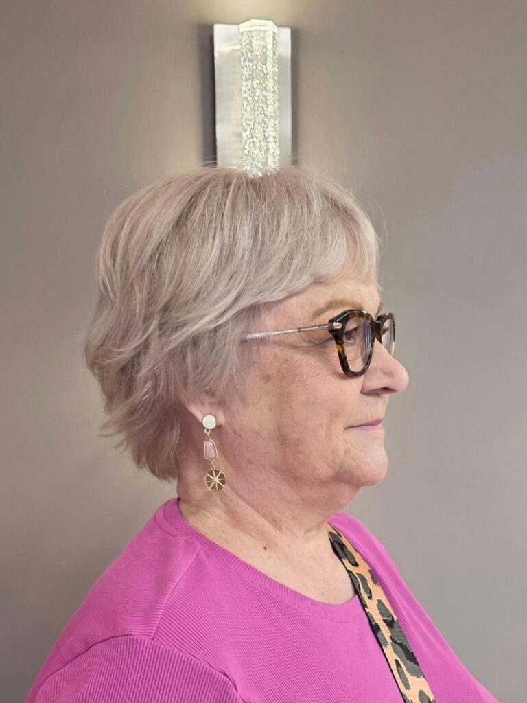A side profile of a woman wearing glasses and showcasing a soft rose blonde feathered bixie haircut with wispy layers.