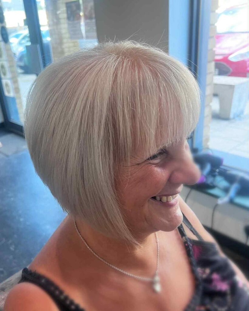 A smiling woman with straight icy white hair is shown in a side view wearing an inverted bob with a full fringe.