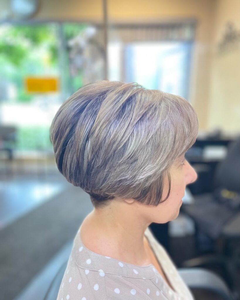 A woman with straight hair is shown from the side wearing a stacked ash blonde bob with subtle grey lowlights and voluminous layers.