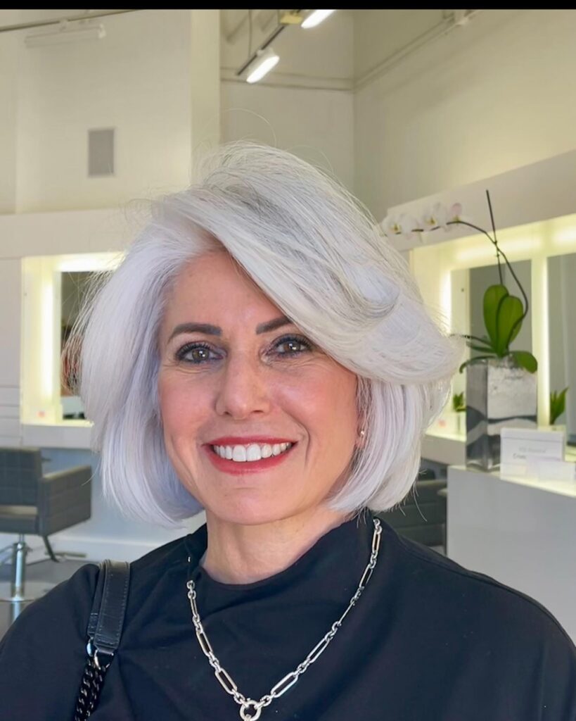 A smiling woman with straight bright silver hair is shown in a three quarter view wearing a voluminous chin length bob with a deep side part and swooping layers.