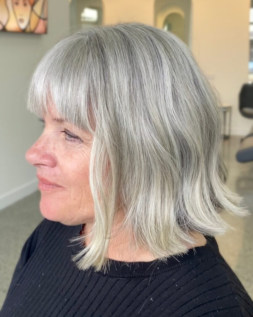 A woman with straight silver hair is shown in a side profile wearing a shoulder length lob with full front bangs.