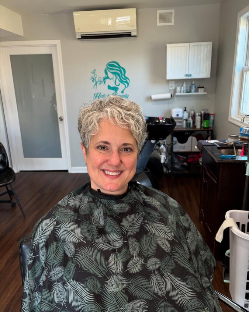 A smiling woman in a salon chair showcasing a short salt and pepper tapered pixie haircut with soft curls styled forward.