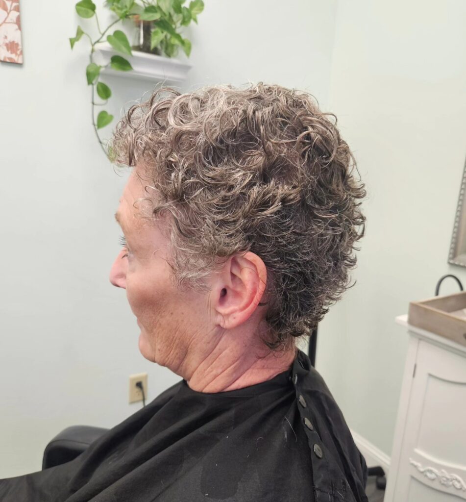 A side profile view of a woman with a salt and pepper tapered pixie haircut featuring damp, finely textured curls.