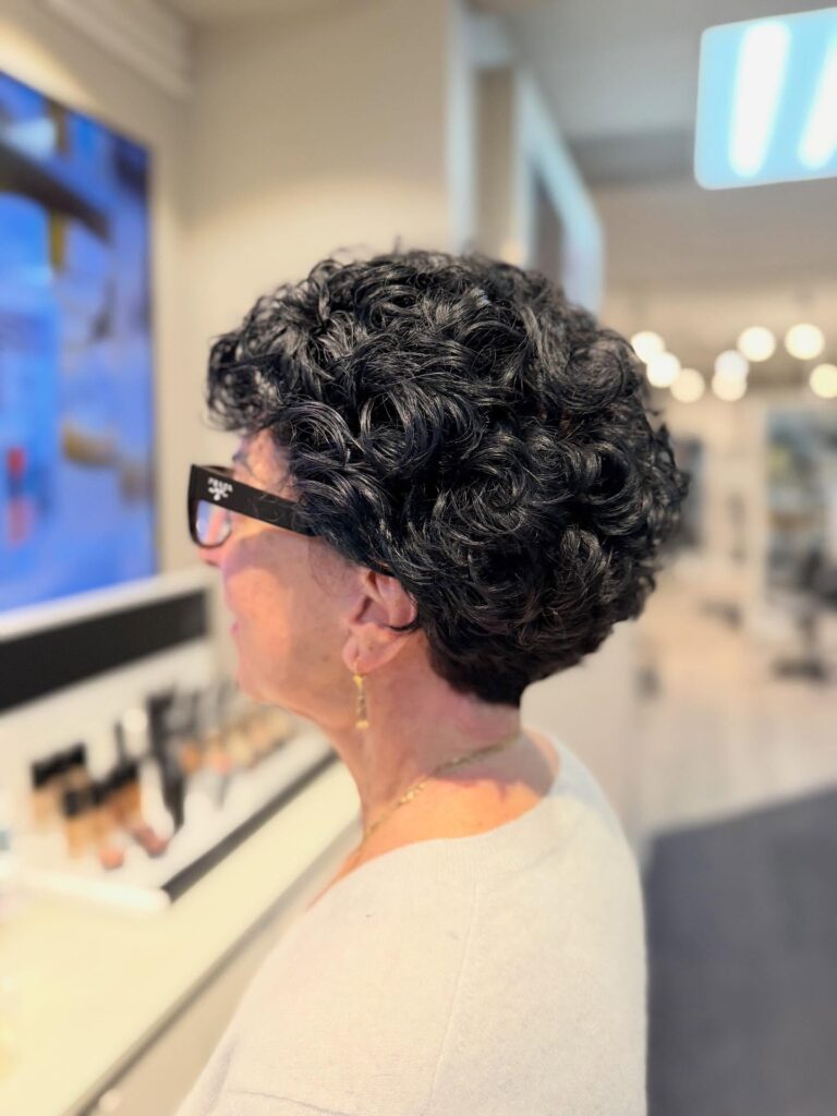 A side profile view of a woman wearing a glossy jet black tapered short haircut with bouncy curls on top.