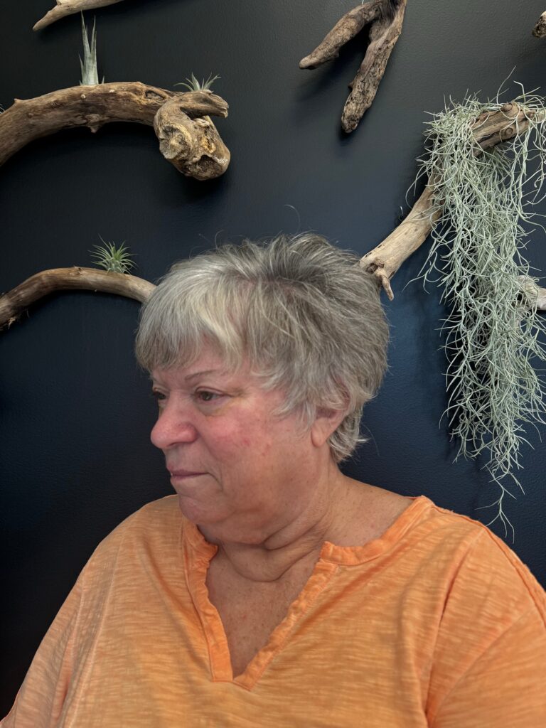 A woman with a short silver and grey pixie cut with wispy bangs stands in profile against a dark wall decorated with driftwood.