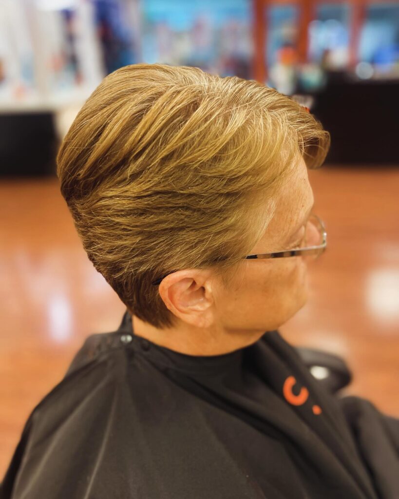 An older woman with glasses smiles, showcasing a short choppy platinum pixie cut with dark roots and texturized layers.