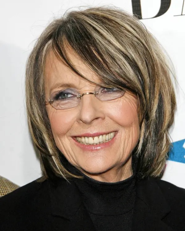 An older woman wearing glasses and a black turtleneck smiles, showcasing a neck length layered bob with ash blonde highlights and a side swept fringe.