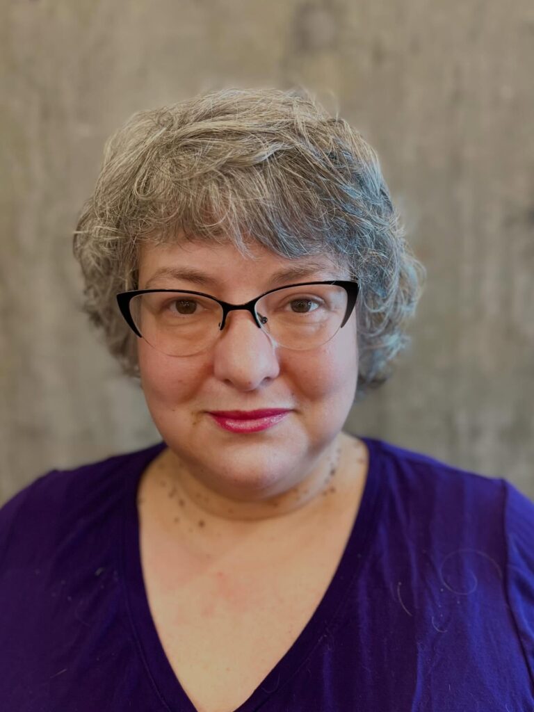 A woman with a short, feathered, ash-gray pixie cut featuring a wispy fringe and soft, wavy texture.