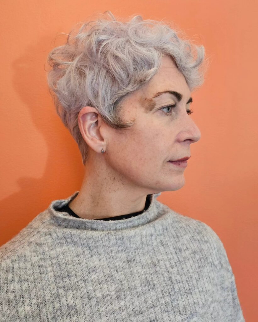 A woman with a short lavender-silver pixie cut featuring voluminous curls on top and tapered sides.