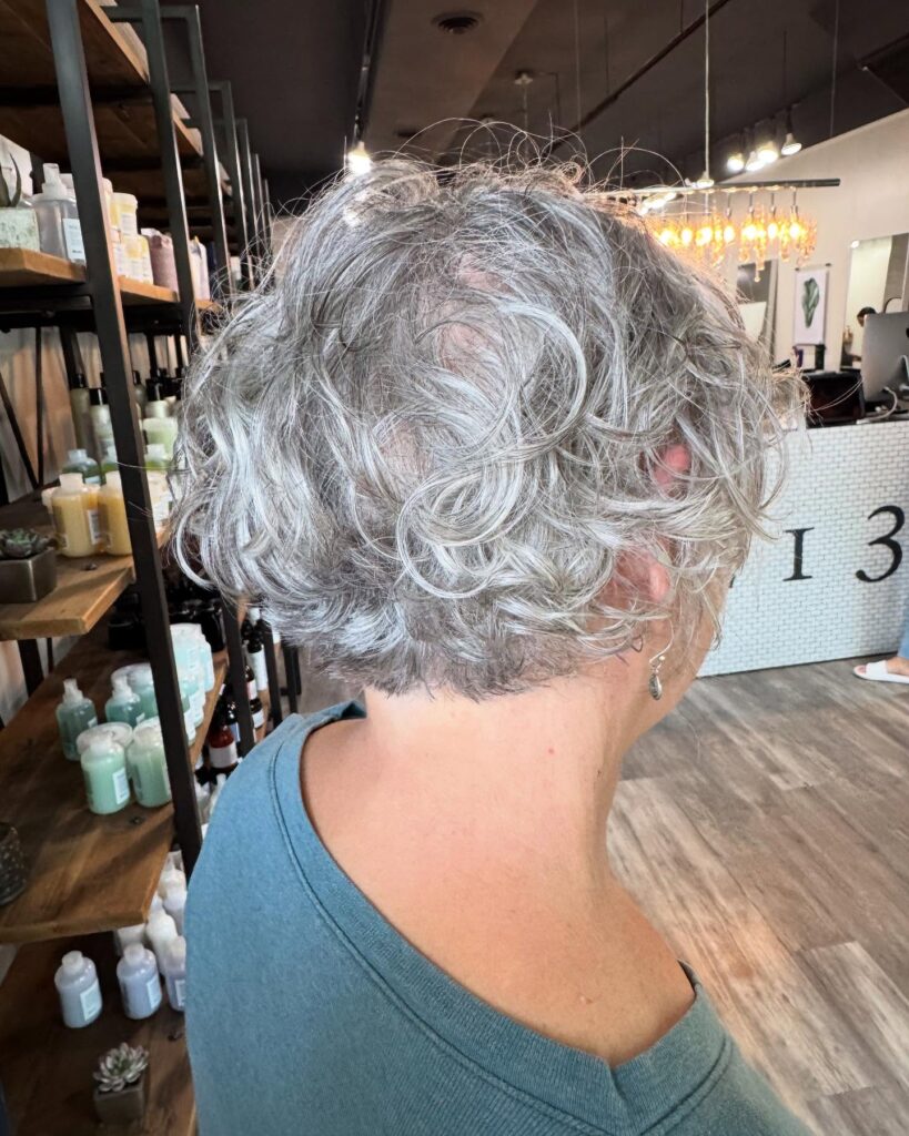 A back view of a woman over 60 with a short, stacked wedge haircut featuring loose grey curls and a tapered neck.