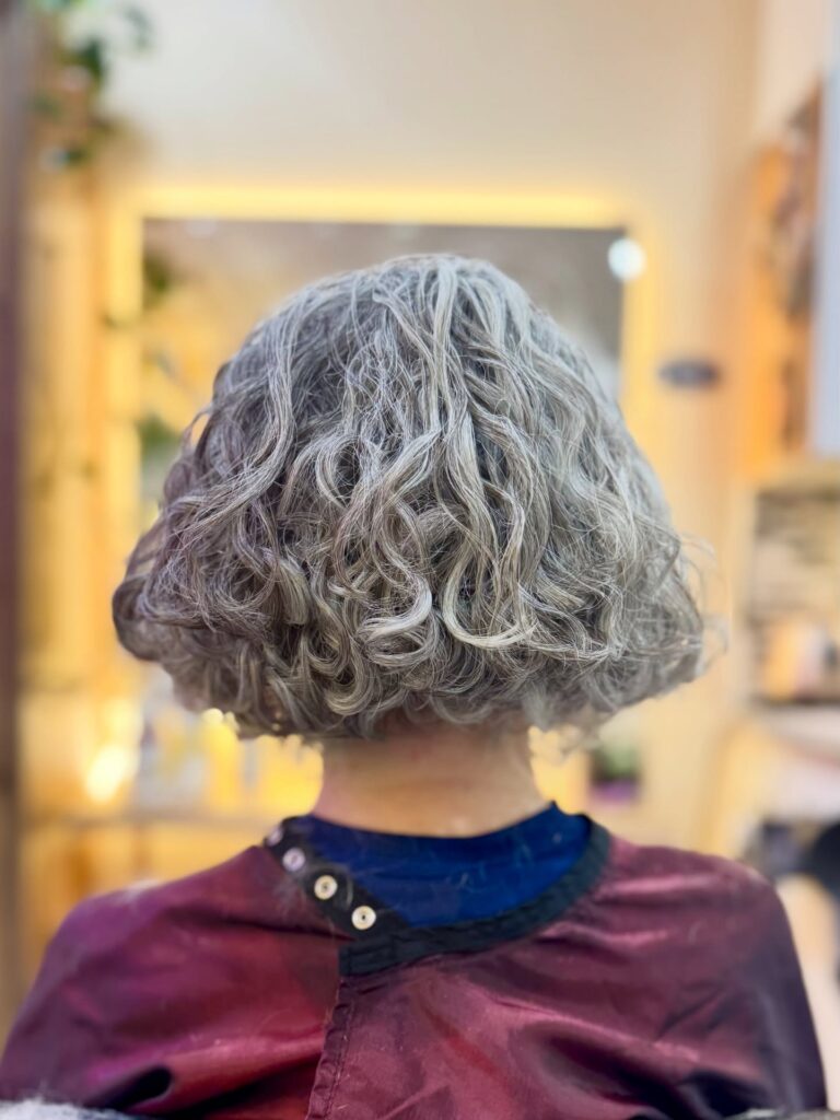 A back view of a woman with a jaw-length French bob featuring loose ash-grey waves and a blunt cut.