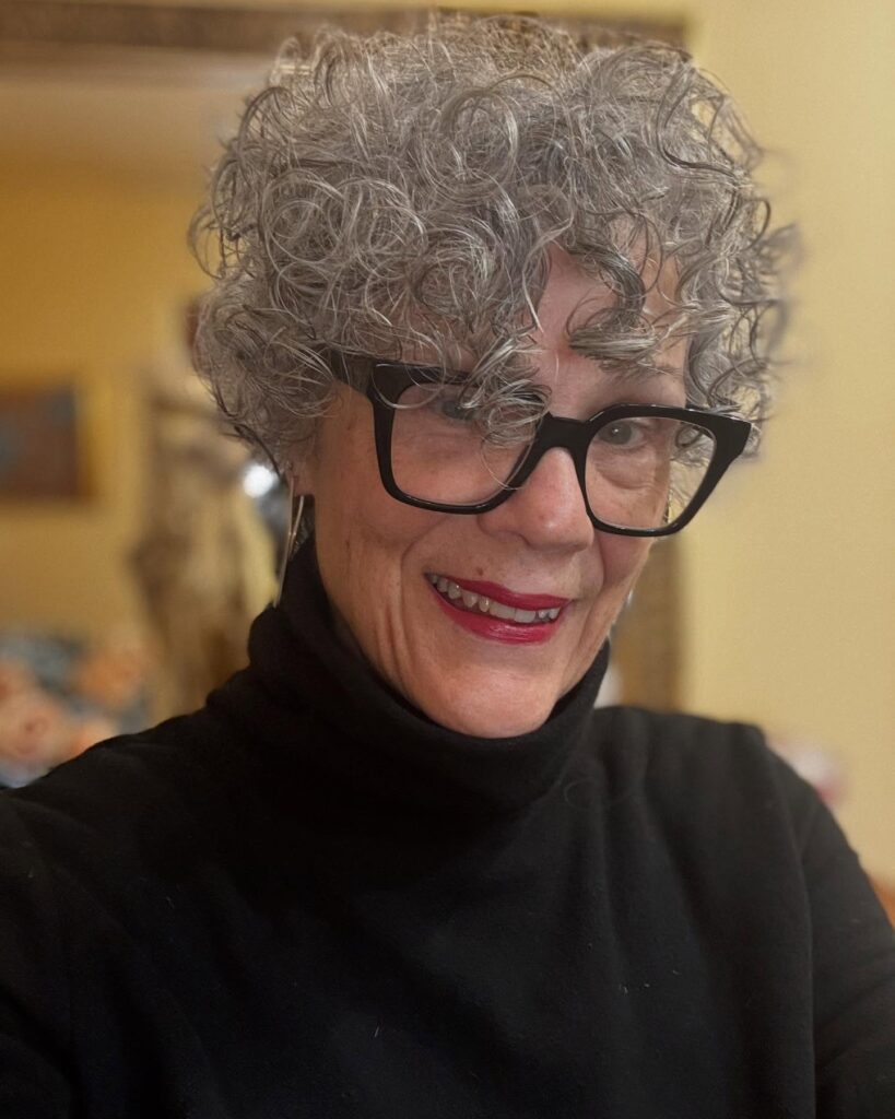A woman wearing black glasses with a short, voluminous charcoal grey hairstyle featuring tight natural ringlets and piecey layers.