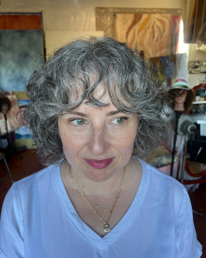 A woman with a short salt-and-pepper curly bob featuring soft layers and curly bangs that frame her face.