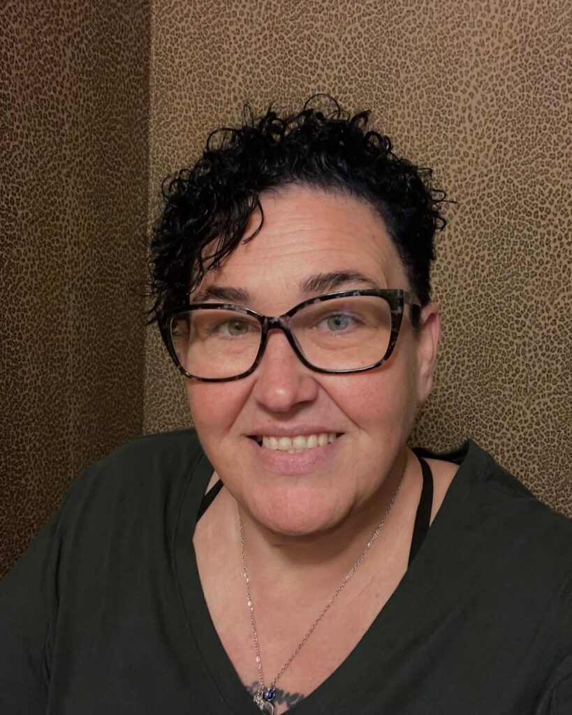 A woman wearing glasses with a short asymmetrical pixie cut featuring tight black curls and closely cropped sides.