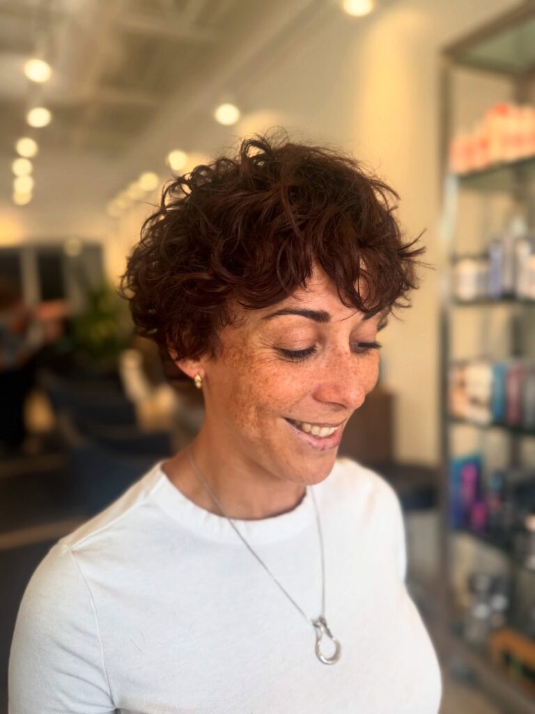 A woman with a short, tousled auburn pixie cut featuring messy curly layers and a soft fringe.