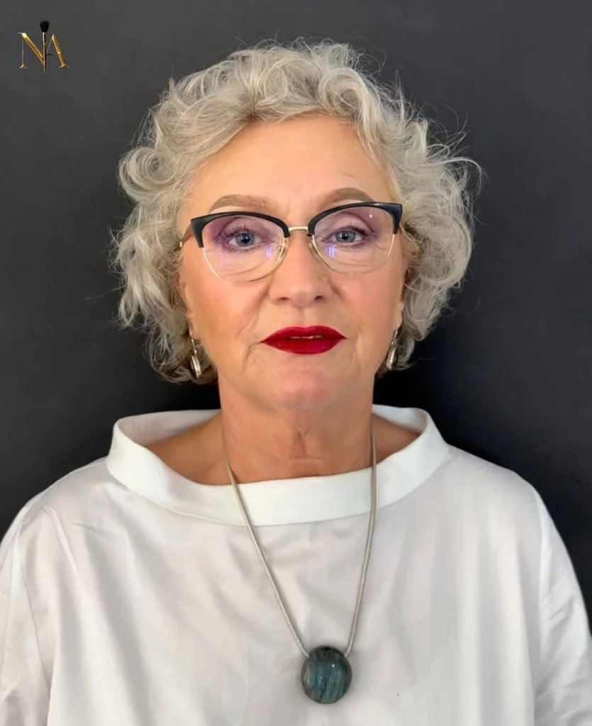 A woman with short curly grey hair styled in a voluminous tousled crop wearing stylish black and gold glasses.