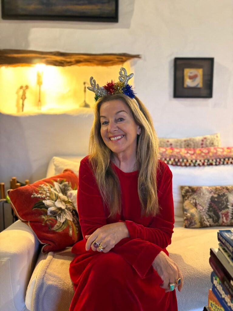 A smiling woman over 60 with long wavy honey blonde hair and a festive headband sits on a sofa.