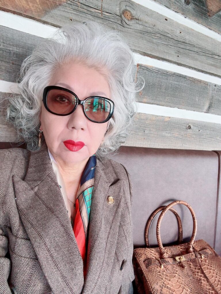 An elegant woman over 60 with voluminous curly silver hair and red lipstick poses in a brown blazer.
