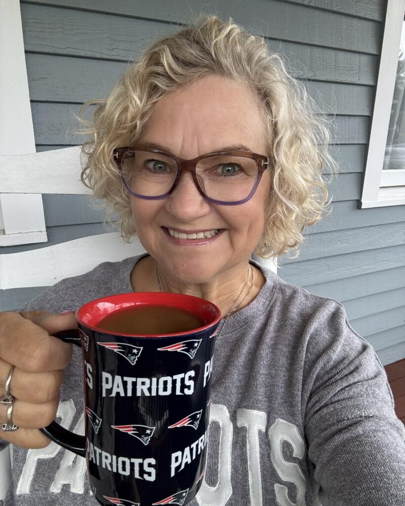 A smiling woman with a chin-length curly blonde bob holding a coffee mug.