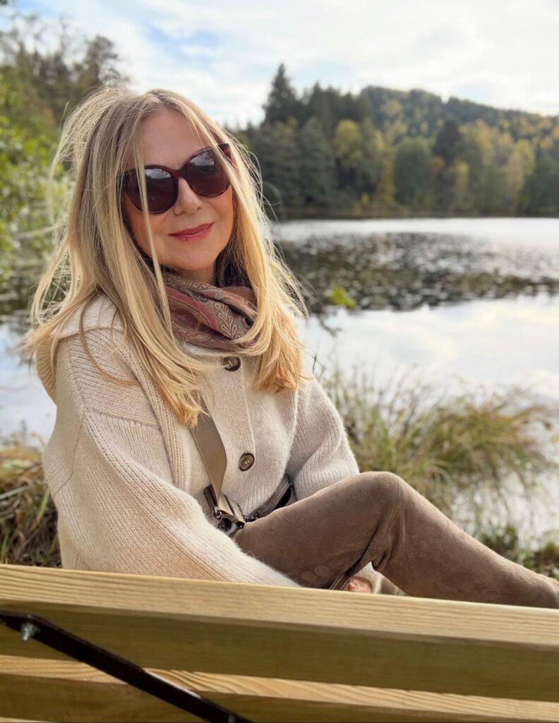 A woman with long sun-kissed blonde hair and soft layers sitting outdoors by a lake.