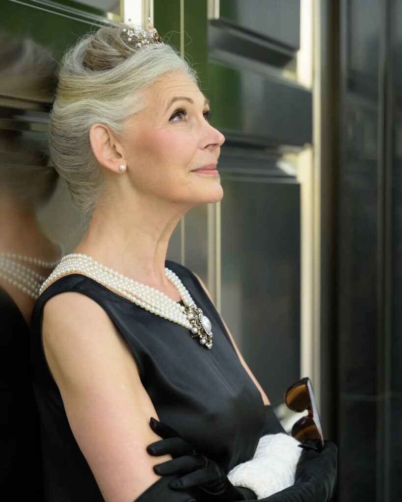 A graceful older woman with a high-volume grey French twist updo adorned with a small pearl and rhinestone tiara.