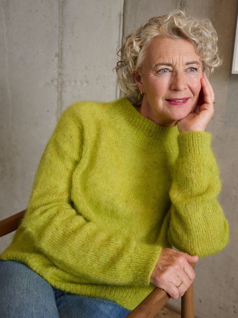 A smiling older woman with a short, bouncy blonde and silver curly pixie hairstyle is wearing a bright green sweater.