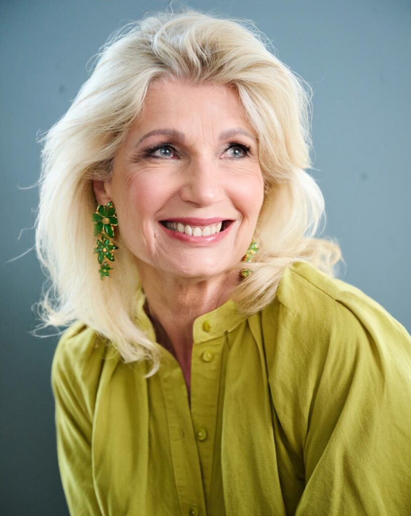 A smiling woman over 60 with a voluminous, mid-length butter blonde hairstyle featuring feathered layers swept back from her face.
