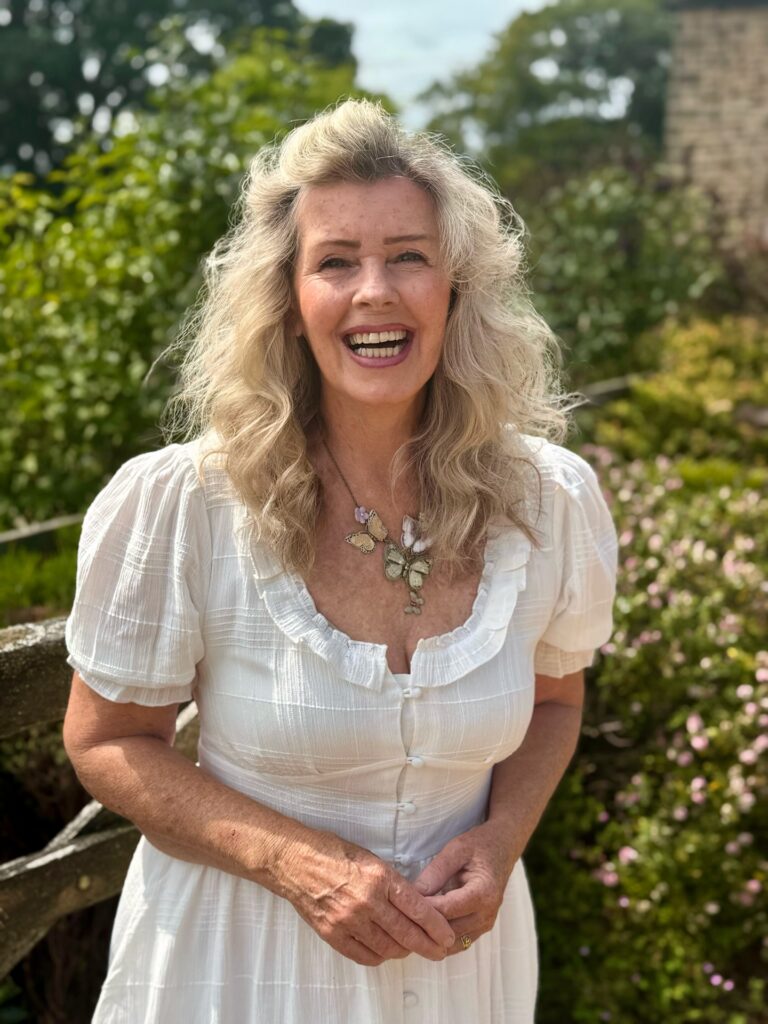 A smiling woman over 60 with long, voluminous blonde hair styled with a swept-back crown and wavy texture.